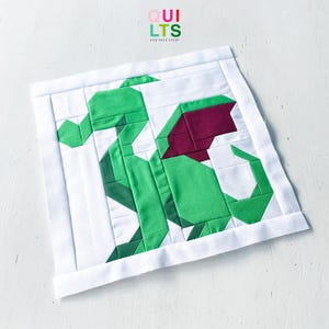 Può includere: Un blocco di trapunta in tessuto quadrato con un design a pezzi di un drago verde con ali rosso scuro su sfondo bianco. Il drago è costruito con segmenti di tessuto geometrici. Il testo QUILTS AND NICE STUFF è visibile sopra.