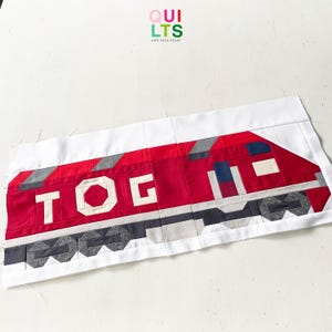 Könnte beinhalten: Ein rot-weißes Quilt-Zug-Design mit den Buchstaben "TOG" in Weiß. Der Zug hat graue und schwarze Akzente und ist vor einem weißen Hintergrund platziert. Im oberen rechten Eck steht der Text "QUILTS AND NICE STUFF".