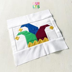 May include: A square fabric quilt block featuring a central jester hat design in blue, green, and maroon with yellow accents. The design is framed by white fabric borders. The text QUILTS AND NICE STUFF is printed at the top center.