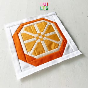 Könnte beinhalten: Ein quadratischer Quiltblock mit einem geometrischen Design in Orange, Gelb und Weiß. Das zentrale Design ist ein Sternmuster, umgeben von einem orangefarbenen Achteck und einem weißen Rand. Der Text "QUILTS AND NICE STUFF" befindet sich oben rechts.