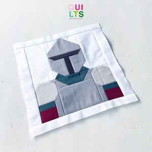 May include: A square fabric quilt block featuring a pieced design of a helmeted figure in shades of gray, teal, and maroon on a white background. The text "QUILTS AND NICE STUFF" is printed in small colorful letters at the top center of the image.