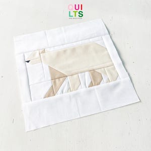 May include: A handmade quilt block featuring a geometric design of a polar bear. The bear is constructed from various shades of beige and white fabric, with a black eye. The block is framed by a white border. The text "QUILTS AND NICE STUFF" is at the top.