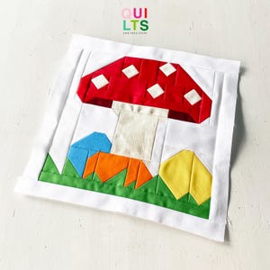 May include: A fabric art piece with a mushroom design. The mushroom has a red cap with white diamond accents, a beige stem, and sits above green, orange, blue, and yellow shapes. The text "QUILTS AND NICE STUFF" is in the upper right corner.