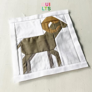 Könnte beinhalten: Ein brauner und beigefarbener geometrischer Patchwork-Quiltblock, der einen Widder darstellt. Der Block ist auf einem weißen Hintergrund genäht. Der Text "OUI LTSS AND NICE STUFF" ist in bunten Buchstaben oben im Bild gedruckt.