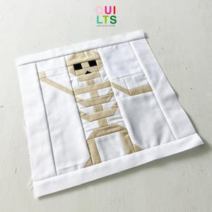 Könnte beinhalten: Ein quadratischer Quiltblock mit einem Mumien-Design. Die Mumie ist beige mit schwarzen Augen und vor einem weißen Hintergrund. Die Worte "QUILTS AND NICE STUFF" befinden sich in der oberen rechten Ecke.