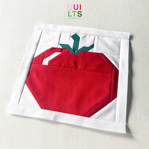 Könnte beinhalten: Ein handgefertigter Quiltblock mit einem geometrischen roten Tomatenmuster, einem grünen Stiel und weißen Akzenten. Die Tomate befindet sich vor einem weißen Hintergrund, der von einem weißen Rand umgeben ist. Der Text "QUILTS" und "AND NICE STUFF" ist sichtbar.