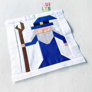 May include: A square fabric quilt block featuring a pieced design of a wizard figure in a blue robe and hat, holding a brown staff. The figure has a light peach face and a grey beard, set against a white background with white borders.