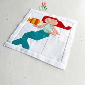 Può includere: Un blocco di patchwork colorato con una sirena con capelli rossi, una coda turchese e un pesce giallo. Il blocco di patchwork è realizzato con diverse tonalità di rosso, arancione, giallo, verde e turchese.