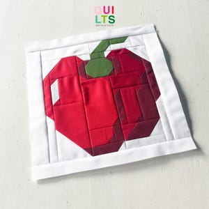 Könnte beinhalten: Ein handgefertigter Quiltblock mit einem roten Apfel mit grünem Stiel und Blatt, vor weißem Hintergrund. Der Apfel besteht aus verschiedenen Rottönen, die ein Patchwork-Design zeigen. Der Text "QUILTS AND NICE STUFF" befindet sich oben rechts.