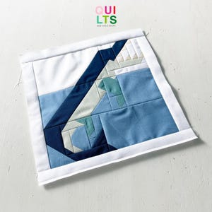 Puede incluir: Un bloque de edredón cuadrado hecho a mano con un diseño geométrico en tonos de azul, blanco y beige. El bloque está bordeado con tela blanca y tiene el texto "QUILTS AND NICE STUFF" en la esquina superior derecha.