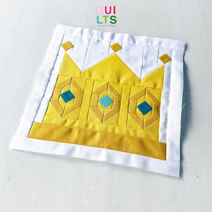 Puede incluir: Un bloque de edredón de tela cuadrado con un diseño de corona amarilla sobre un fondo blanco. La corona está decorada con tres detalles en forma de diamante de color azul verdoso y pequeños cuadrados amarillos en las puntas. El texto QUILTS AND NICE STUFF es visible.