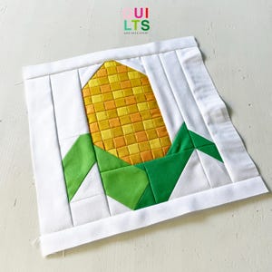 May include: A square fabric quilt block featuring a central corn cob design made of yellow and orange squares, surrounded by green fabric leaves. The entire design is framed by white fabric borders, with the text QUILTS AND NICE STUFF above.