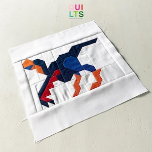 Peut inclure: Un carré de tissu blanc avec un motif géométrique d'un oiseau dans des tons de bleu, rouge et orange. Le motif est composé de différentes formes et tailles de morceaux de tissu cousus ensemble. Le carré de tissu est plié en deux et les bords sont visibles.