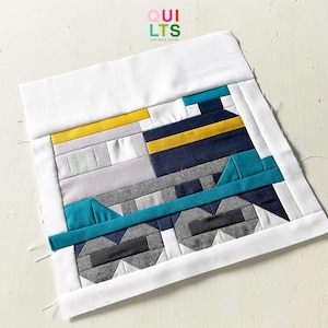 Könnte beinhalten: Ein farbenfroher Quiltblock mit geometrischen Formen in Blau-, Grau-, Gelb- und Türkistönen. Der Block ist mit einem weißen Rand versehen und trägt den Text "QUILTS AND NICE STUFF" in der oberen rechten Ecke.