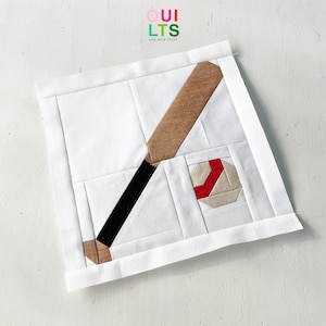 Puede incluir: Un bloque de colcha de tela blanca con un bate de béisbol marrón y negro y una pelota de béisbol roja y blanca. El bloque de colcha está rodeado por un borde blanco.