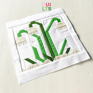 May include: A white and green quilted block featuring a lily of the valley design. The quilt block has a white background with green stems and leaves, and off-white bell-shaped flowers. The text "QUILTS AND NICE STUFF" is in the upper right corner.