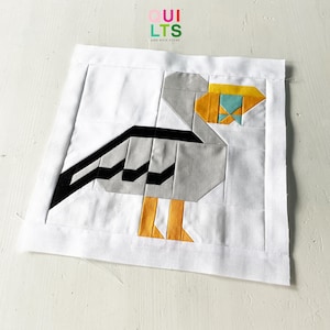 Könnte beinhalten: Ein weißer Stoffquadrat mit einem geometrischen Vogeldesign in Grau-, Schwarz- und Gelbtönen. Der Vogel besteht aus zusammengenähten Stoffstücken. Der Stoffquadrat ist von einem weißen Rand umgeben.