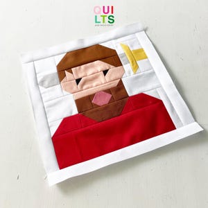 May include: A handmade quilt block featuring a geometric design of a person with brown hair and beard, wearing a red shirt. The block is framed in white fabric and includes the text "QUILTS AND NICE STUFF" in the upper left corner.