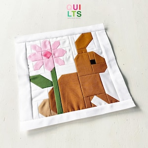 Könnte beinhalten: Ein handgefertigter Quiltblock mit einem braunen Hasen und einer rosa Blume. Der Hase und die Blume bestehen aus geometrischen Formen, vor einem weißen Hintergrund. Der Block ist mit einem weißen Rand versehen. Der Text "QUILTS and NICE STUFF" ist sichtbar.