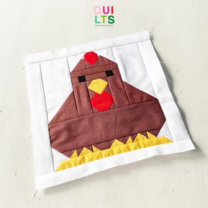 May include: A patchwork fabric block depicting a stylised chicken. The chicken is brown with red and yellow details, set on a white background. The text "QUILTS AND NICE STUFF" is visible in the upper left corner.