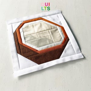 Könnte beinhalten: Ein quadratischer Quiltblock mit geometrischem Design. Die Mitte zeigt beigen und grauen Stoff, umgeben von Schichten aus Braun und Orange, alles eingerahmt von einem weißen Rand. Der Text "QUILTS" und "AND NICE STUFF" ist oben rechts sichtbar.