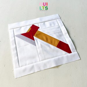 May include: A square fabric quilt block featuring a pieced design of an axe with a grey blade, a red head, and a mustard yellow handle. The axe is set against a solid white background with white fabric borders on all four sides of the square.
