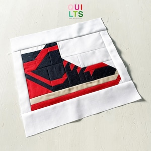 May include: A square fabric quilt block featuring a pieced design of a high-top sneaker in black, red, and beige, framed by a white border. The text QUILTS AND NICE STUFF is printed in small colorful letters at the top center of the composition.