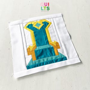Può includere: Un blocco trapuntato quadrato in tessuto con un design centrale a forma di trono in blu ottanio, giallo e azzurro, incorniciato da un bordo bianco. Il design è composto da pezzi di tessuto geometrici cuciti, con il testo QUILTS AND NICE STUFF sopra.