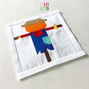 Peut inclure: Un bloc de patchwork représentant un épouvantail. L'épouvantail a un chapeau marron, un visage orange, des bras rouges, une salopette bleue et une tige marron. Le bloc est entouré de tissu blanc et contient le texte "QUILTS AND NICK STUFF".