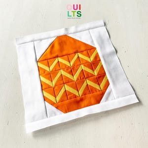 May include: A square quilt block with an orange and yellow geometric pattern, similar to candy corn. The design is set against a white background, with the text "QUILTS AND NICE STUFF" in the upper left corner.