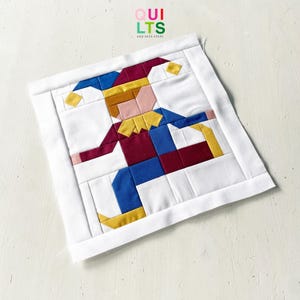 Könnte beinhalten: Ein quadratischer Quiltblock aus Stoff mit einem zusammengesetzten Design einer Narrenfigur in Bewegung. Der Narr besteht aus einfarbigen Stoffblöcken in Weinrot, Blau, Gelb und Hellrosa, umrahmt von einem weißen Rand. Der Text QUILTS AND NICE STUFF ist sichtbar.