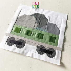 May include: A fabric quilt block featuring a train design. The train is composed of various shades of gray and green fabric, with black wheels. The block is bordered by white fabric. The text "QUILTS AND NICE STUFF" is in the upper right corner.