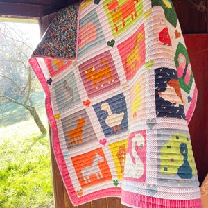 May include: A pink and white patchwork quilt featuring various farm animals, including a duck, a pig, a horse, a cat, and a bird. The quilt is hanging on a wooden door.