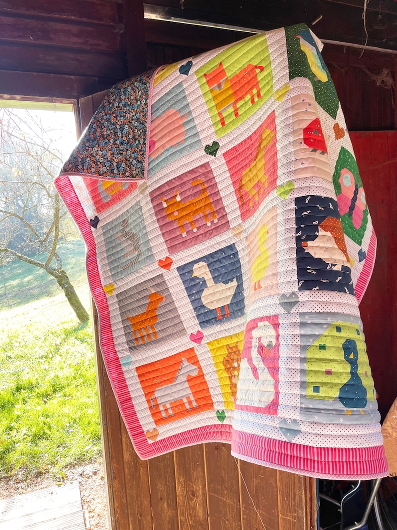 May include: A pink and white patchwork quilt featuring various farm animals, including a duck, a pig, a horse, a cat, and a bird. The quilt is hanging on a wooden door.
