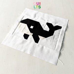 Op de afbeelding: Een wit vierkant van stof met een zwart-wit gepixelde walvisontwerp. De stof heeft een rauwe rand en is aan elkaar gestikt. De tekst "QUI LTS AND NICE STUFF" is in kleurrijke letters bovenaan de stof gedrukt.