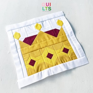 Op de afbeelding: Een vierkant quiltblok met een centraal geel kroonontwerp met dieprode ruitvormige accenten en kleine gele zeshoekige vormen aan de bovenkant. De kroon is omlijst door een witte rand en de tekst QUILTS AND NICE STUFF is erboven zichtbaar.
