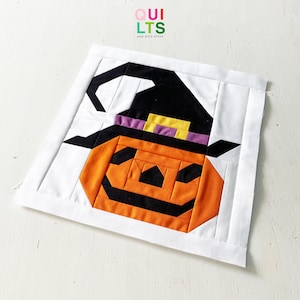 Könnte beinhalten: Ein Halloween-Quiltblock mit einem orangefarbenen Kürbis, der einen schwarzen Hexenhut mit lila und gelben Akzenten trägt. Das Design ist vor einem weißen Hintergrund platziert, mit dem Text "QUILTS AND NICE STUFF" in der oberen linken Ecke.