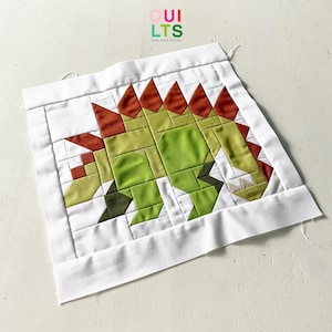 Könnte beinhalten: Ein weißer Stoff-Quiltblock mit einem grünen und roten Patchwork-Design eines Dinosauriers. Der Quiltblock ist mit "QUI LTS AND NICE STUFF" beschriftet.