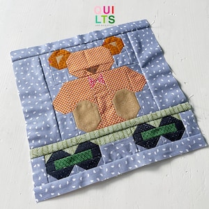 May include: A patchwork quilt block featuring a brown teddy bear with a red bow tie, standing on a green and black patterned base. The block is set against a blue background with white polka dots.