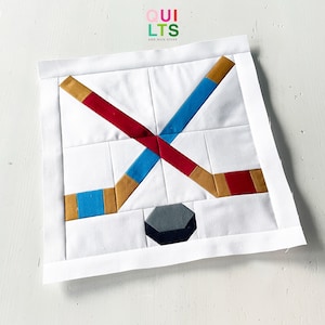 May include: A white square quilt block with a hockey theme. It features a gray and black puck and crossed hockey sticks in red, blue, and gold. The text "QUILTS AND NICE STUFF" is in the upper right corner.