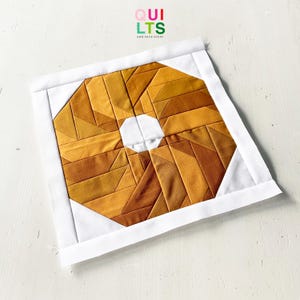 May include: A square quilt block with a geometric pattern in shades of gold and brown, with a white centre and border. The text "QUILTS AND NICE STUFF" is in the upper right corner.