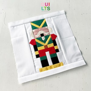 May include: A square fabric block with a pixelated nutcracker design in green, red, yellow, and black. The design is set against a white background. The text "QUILTS AND NICE STUFF" is visible in the upper right corner of the image.