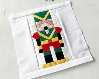May include: A square fabric block featuring a colorful, pixelated nutcracker design. The nutcracker is dressed in green, red, yellow, and black, set against a white background. The text "QUILTS AND NICE STUFF" is in the upper right corner.
