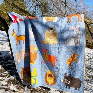 May include: A blue patchwork quilt featuring various animal designs, such as a fox, a cow, a rabbit, and a bear. The quilt is draped over a tree branch in a snowy woodland scene.