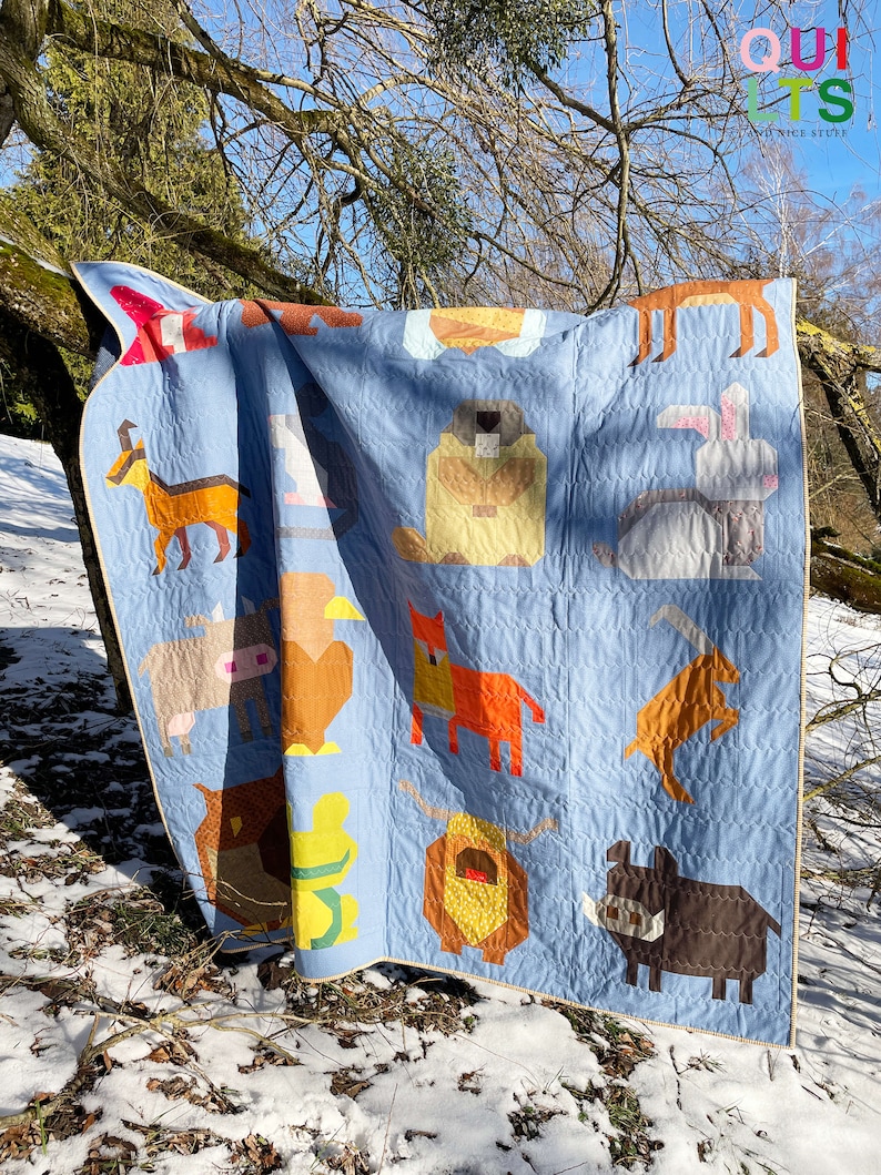 May include: A blue patchwork quilt featuring various animal designs, such as a fox, a cow, a rabbit, and a bear. The quilt is draped over a tree branch in a snowy woodland scene.