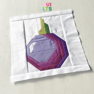 Könnte beinhalten: Ein Stoff-Quiltblock mit einem geometrischen Design einer lila und magenta farbenen Frucht mit grünen und gelben Akzenten. Die Frucht ist vor einem weißen Hintergrund platziert, mit dem Text "Quilts and Nice Stuff" in der oberen rechten Ecke.