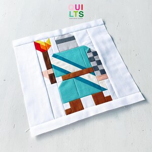 May include: A square fabric quilt block featuring a pieced design of a knight in teal armour holding a torch with a red and yellow flame. The figure has a grey helmet, brown boots, and a brown belt, all set against a solid white background border.