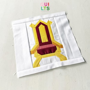 Op de afbeelding: Een vierkant stoffen quiltblok met een centraal samengesteld ontwerp van een stoel in tinten geel, bordeauxrood en roze, omringd door een witte rand. De tekst "QUILTS AND NICE STUFF" staat in kleine gekleurde letters bovenaan in het midden.