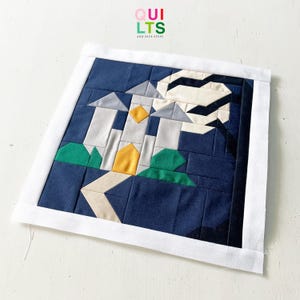 May include: A quilted wall hanging with a geometric castle design under a moonlit sky. The quilt uses blue, grey, yellow, green, and white. The text "QUILTS AND NICE STUFF" is in the upper right corner.