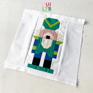 Könnte beinhalten: Ein weißer Stoffquadrat mit einem Patchwork-Design eines Nussknackers in Grün, Blau, Schwarz und Rosa. Der Stoffquadrat hat den Text "QUILTS AND NICE STUFF" in der oberen linken Ecke.